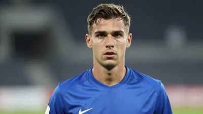 Mid-30s Sandefjord forward, athletic build, intense gaze, head-and-shoulders, wearing a plain blue shirt with white...