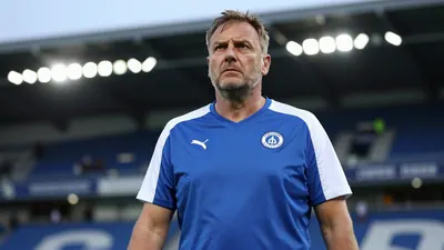 A weary, triumphant Sandefjord coach, mid-40s, stocky build, in a plain blue and white training top, stands pitchside at...
