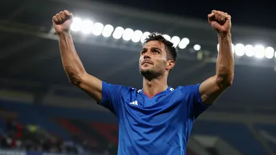 A muscular male footballer in a blue jersey, mid-20s, celebrating a goal with arms raised