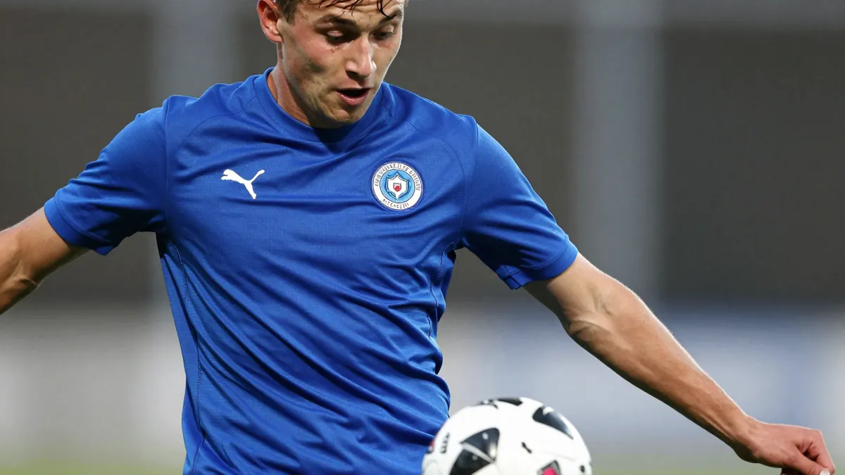 A close-up of an anonymous 18-year-old Sandefjord player in a solid blue kit, mid-action as he strikes a ball, face...