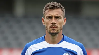 A determined, rugged Sandefjord defender, mid-30s, with a strong build, wearing a plain blue and white training top...